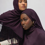 Two women wearing deep plum purple hijabs with fringe trim, posed closely against a plain light gray background.