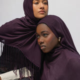 Two women wearing deep plum purple hijabs with fringe trim, posed closely against a plain light gray background.