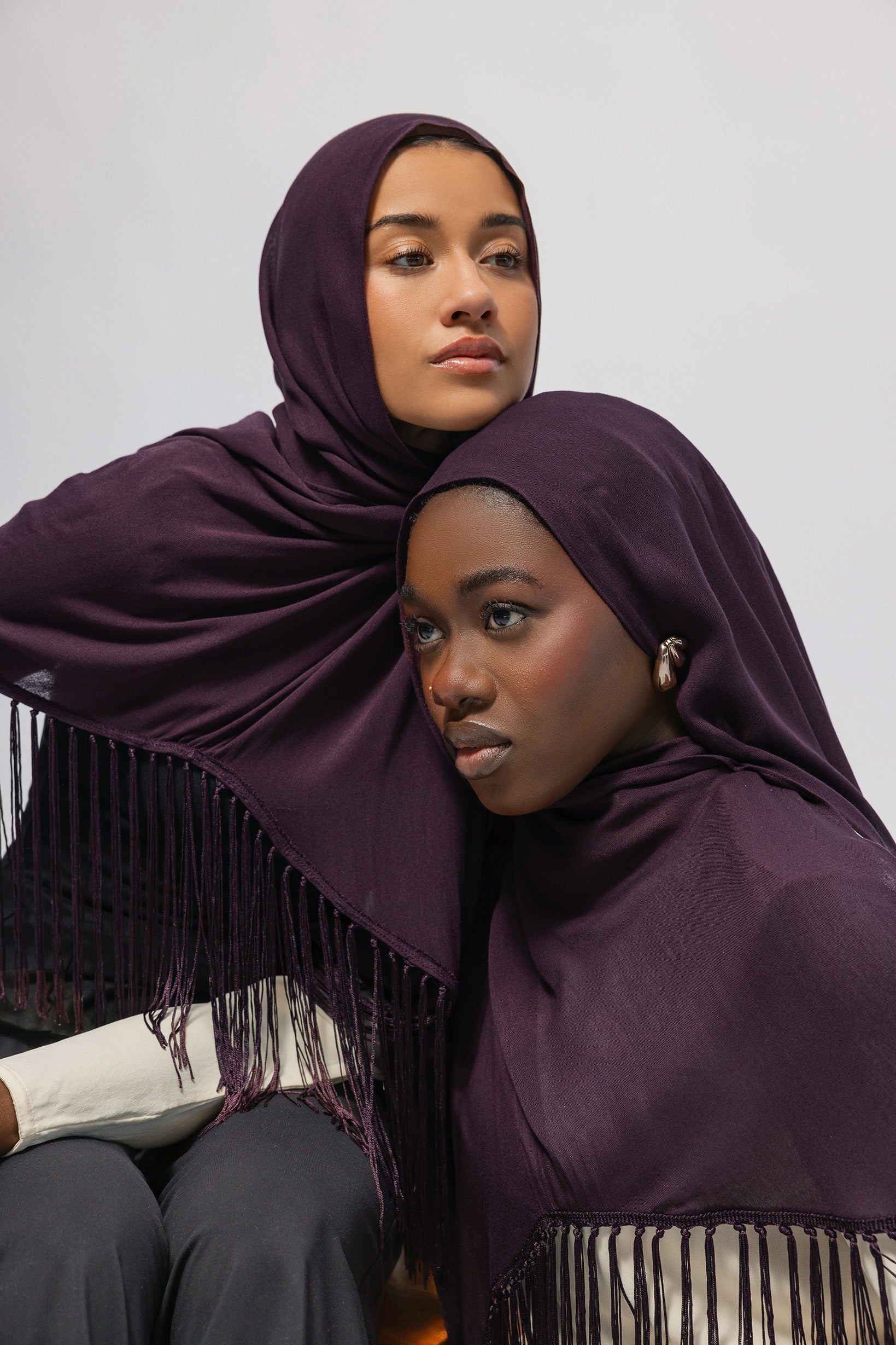 Two women wearing deep plum purple hijabs with fringe trim, posed closely against a plain light gray background.
