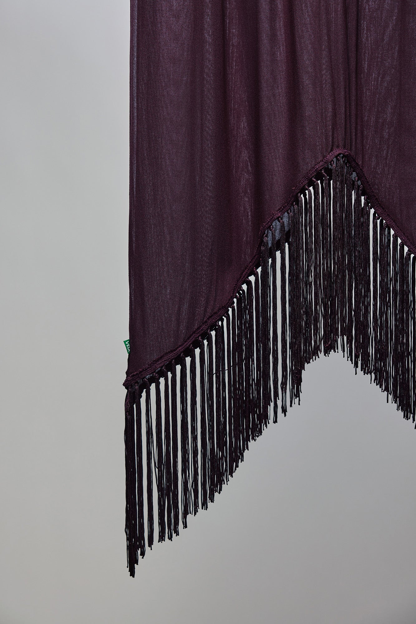 A dark plum purple fabric panel with long black fringe trim along the jagged curved bottom edge, shown against a plain light gray background.