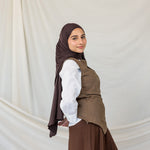 Woman wearing a brown hijab, white shirt, and brown vest and skirt against a plain background