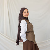 Woman wearing a brown hijab, white shirt, and brown vest and skirt against a plain background