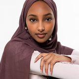A woman wearing a draped plum-colored hijab and a light pink top, leaning on a black chair against a plain white background.