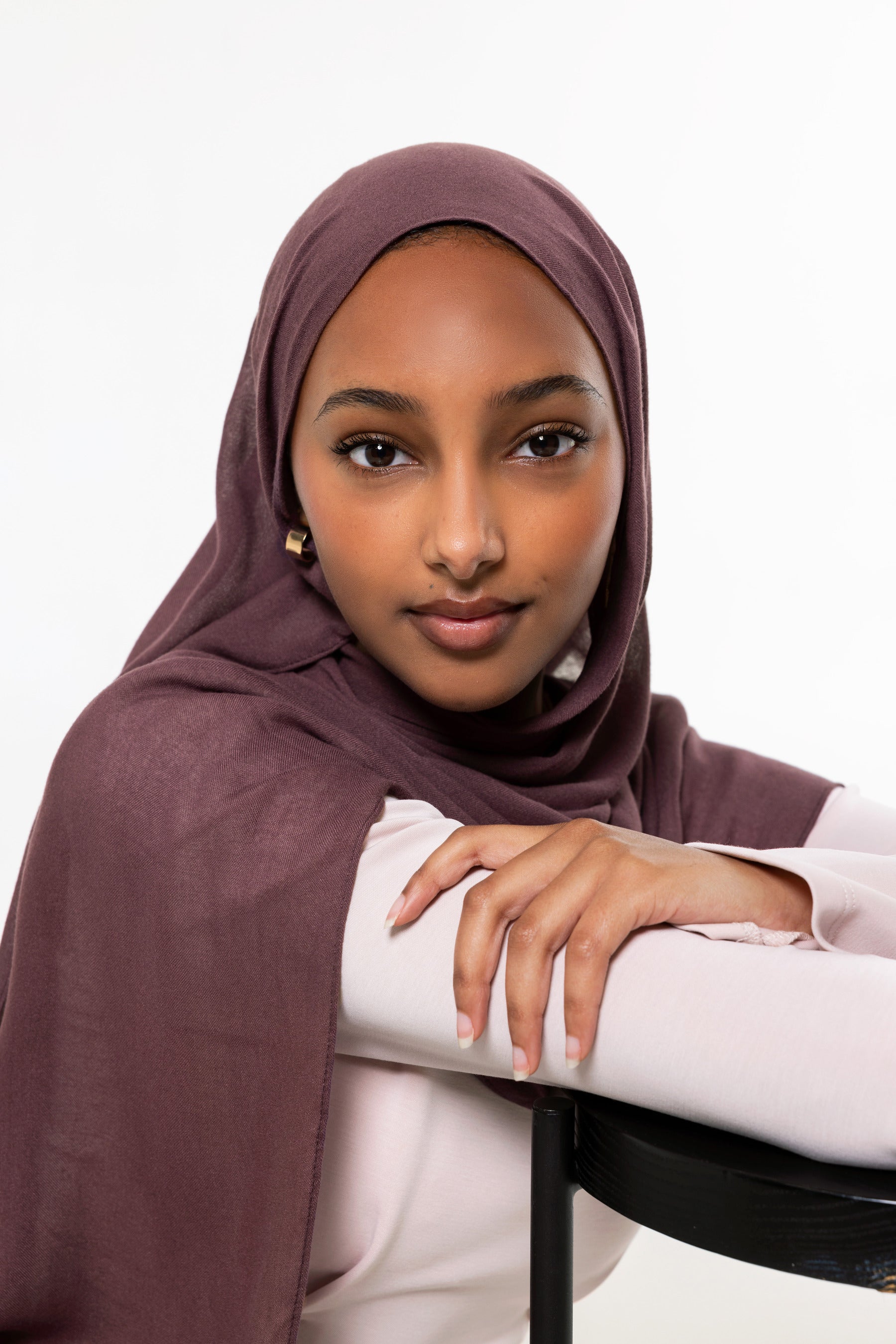 A woman wearing a draped plum-colored hijab and a light pink top, leaning on a black chair against a plain white background.