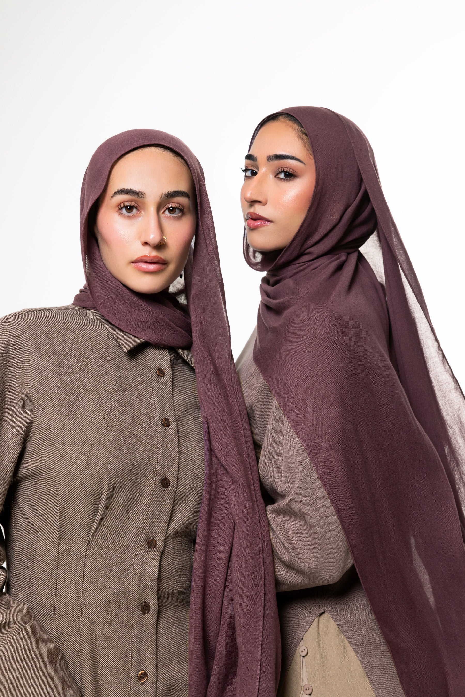 Two women modeling matching plum-colored hijabs and coordinated taupe button-front tops against a plain light gray studio background.