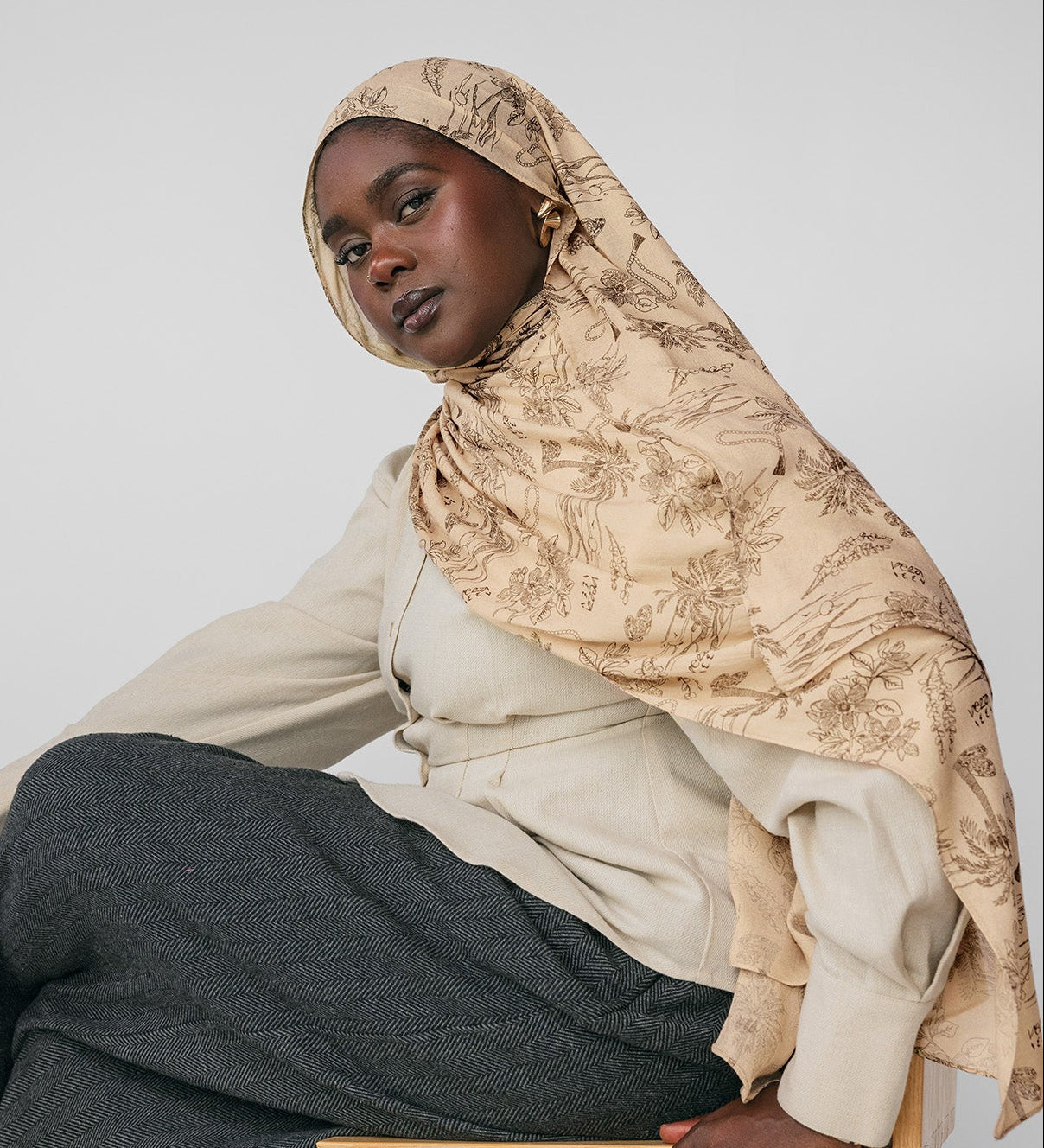 Woman wearing a beige hijab and gray pants sitting on a wooden stool against a light gray background