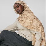 Woman wearing a beige hijab and gray pants sitting on a wooden stool against a light gray background