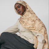 Woman wearing a beige hijab and gray pants sitting on a wooden stool against a light gray background