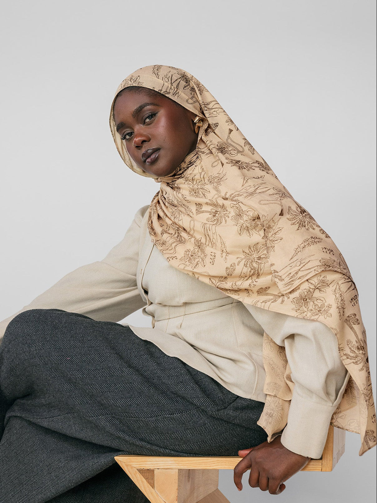 Woman wearing a beige hijab and gray pants sitting on a wooden stool against a light gray background