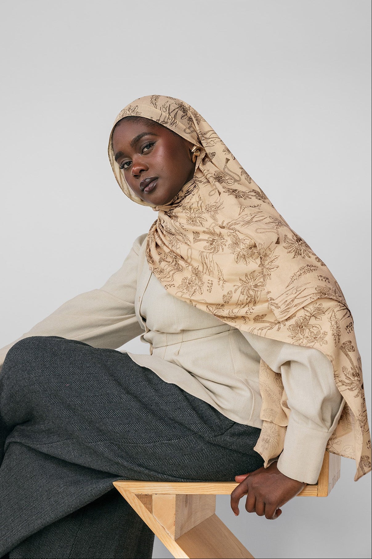 Woman wearing a beige hijab and gray pants sitting on a wooden stool against a light gray background
