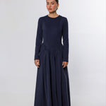 A woman modeling a long-sleeved navy blue maxi dress with a gathered waist, standing against a plain light gray studio background.