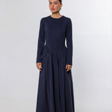 A woman modeling a long-sleeved navy blue maxi dress with a gathered waist, standing against a plain light gray studio background.