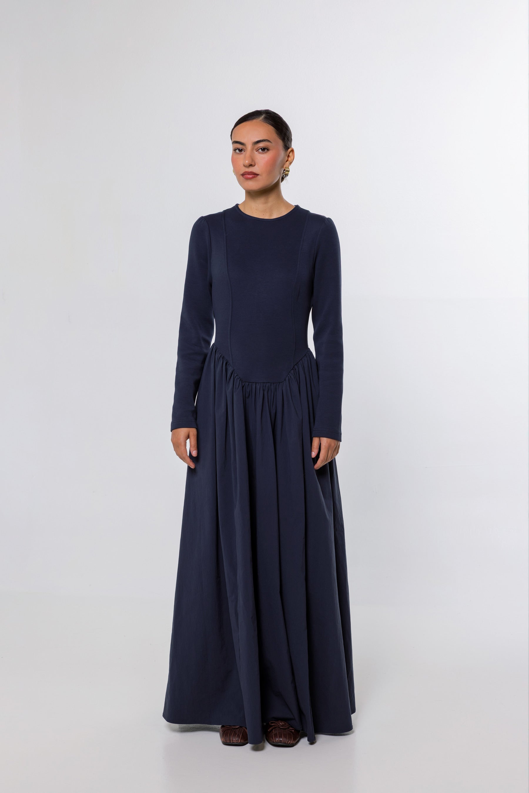 A woman modeling a long-sleeved navy blue maxi dress with a gathered waist, standing against a plain light gray studio background.
