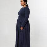 A woman modeling a long-sleeved navy blue maxi dress, shown in profile against a plain light gray studio background.