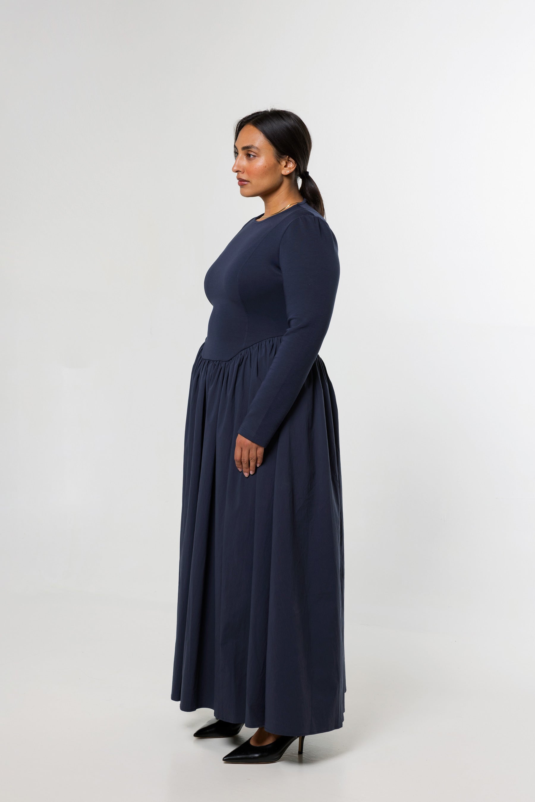 A woman modeling a long-sleeved navy blue maxi dress, shown in profile against a plain light gray studio background.