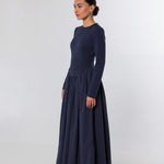 A woman modeling a long-sleeved navy blue maxi dress with a fitted bodice and a full, flowing skirt, photographed against a light gray studio background.