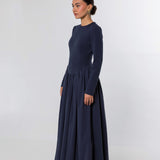 A woman modeling a long-sleeved navy blue maxi dress with a fitted bodice and a full, flowing skirt, photographed against a light gray studio background.