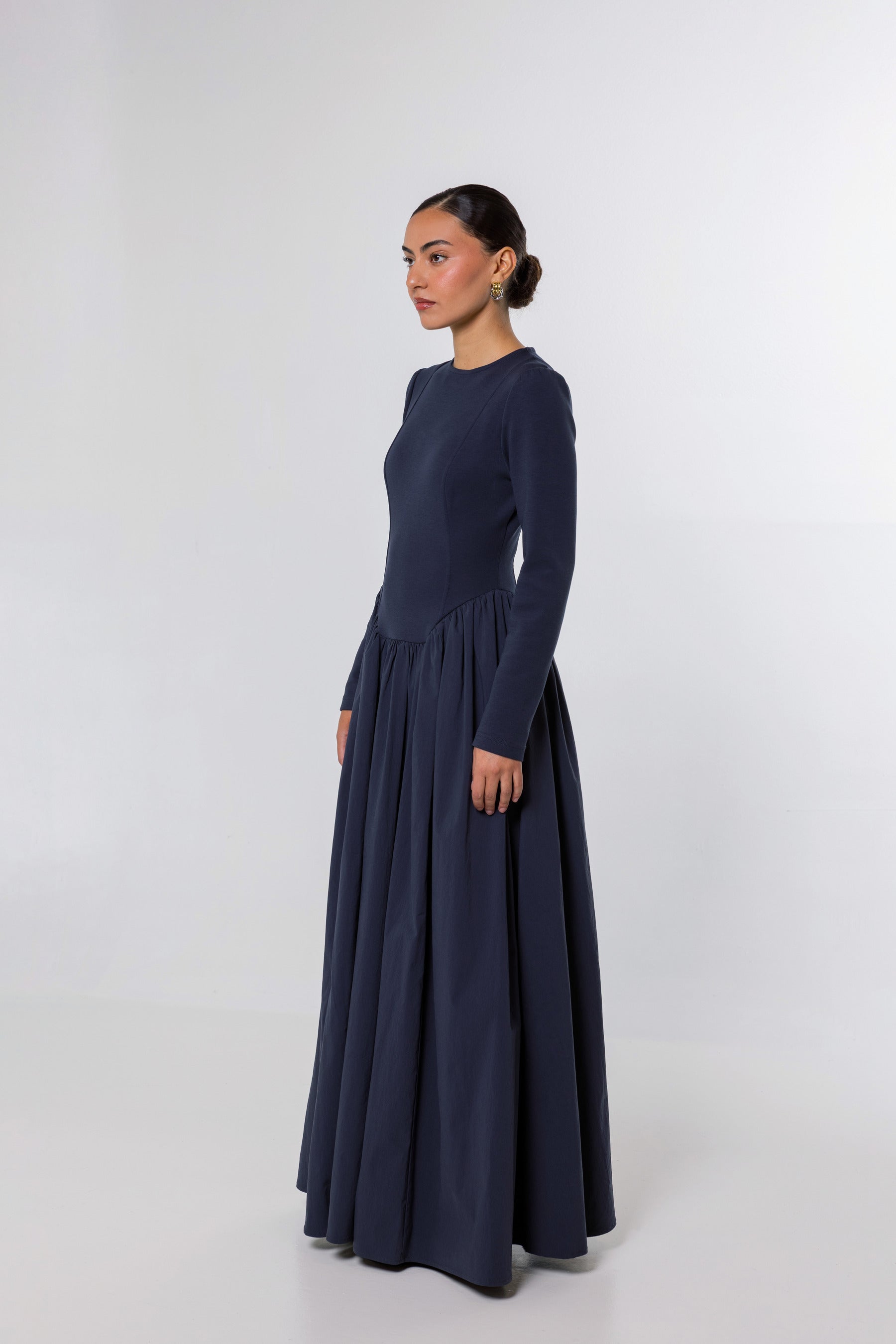 A woman modeling a long-sleeved navy blue maxi dress with a fitted bodice and a full, flowing skirt, photographed against a light gray studio background.