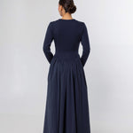 A woman viewed from the back wearing a long-sleeve navy maxi dress with a fitted bodice and a full gathered skirt, standing against a plain light gray studio background.