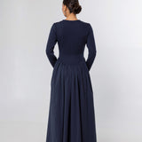 A woman viewed from the back wearing a long-sleeve navy maxi dress with a fitted bodice and a full gathered skirt, standing against a plain light gray studio background.