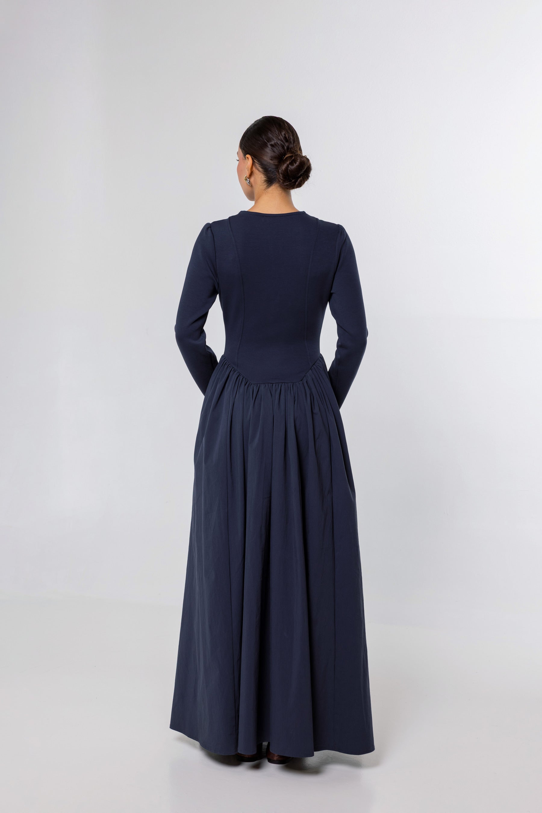 A woman viewed from the back wearing a long-sleeve navy maxi dress with a fitted bodice and a full gathered skirt, standing against a plain light gray studio background.