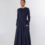 A woman modeling a long-sleeved navy blue maxi dress with a gathered waist and side pockets, standing against a light gray studio background.