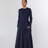 A woman modeling a long-sleeved navy blue maxi dress with a gathered waist and side pockets, standing against a light gray studio background.