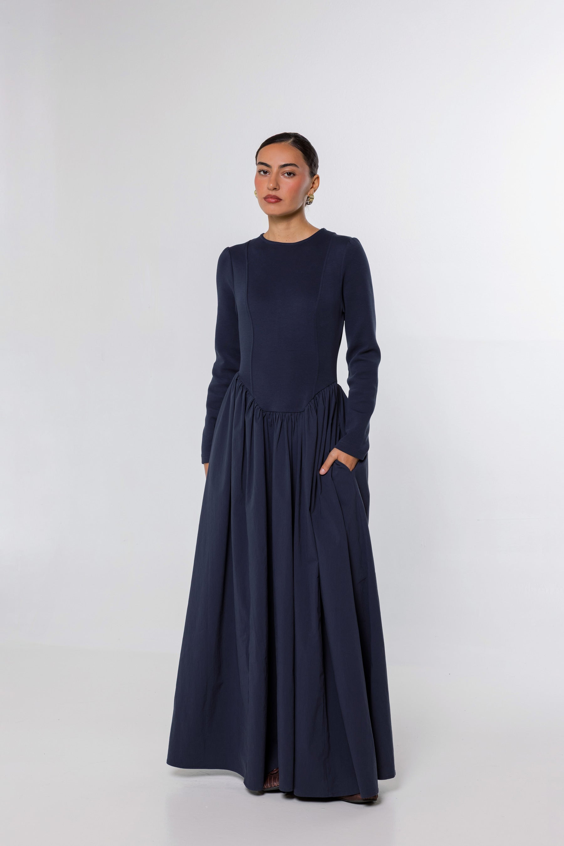 A woman modeling a long-sleeved navy blue maxi dress with a gathered waist and side pockets, standing against a light gray studio background.