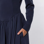 Close-up of a navy blue long-sleeve dress with a gathered waist and side pocket, shown on a model with one hand in the pocket against a light gray background.