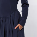 Close-up of a navy blue long-sleeve dress with a gathered waist and side pocket, shown on a model with one hand in the pocket against a light gray background.