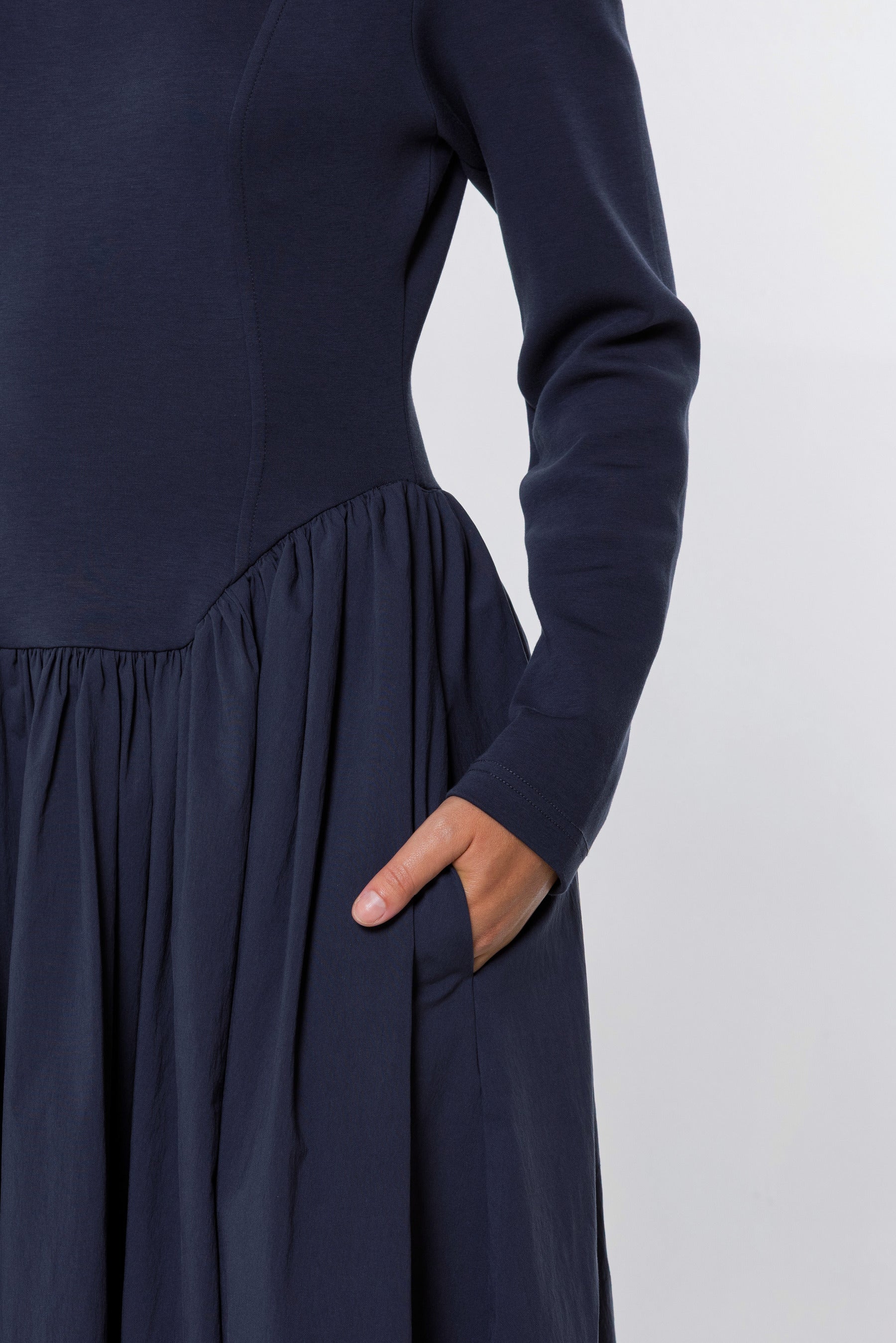 Close-up of a navy blue long-sleeve dress with a gathered waist and side pocket, shown on a model with one hand in the pocket against a light gray background.