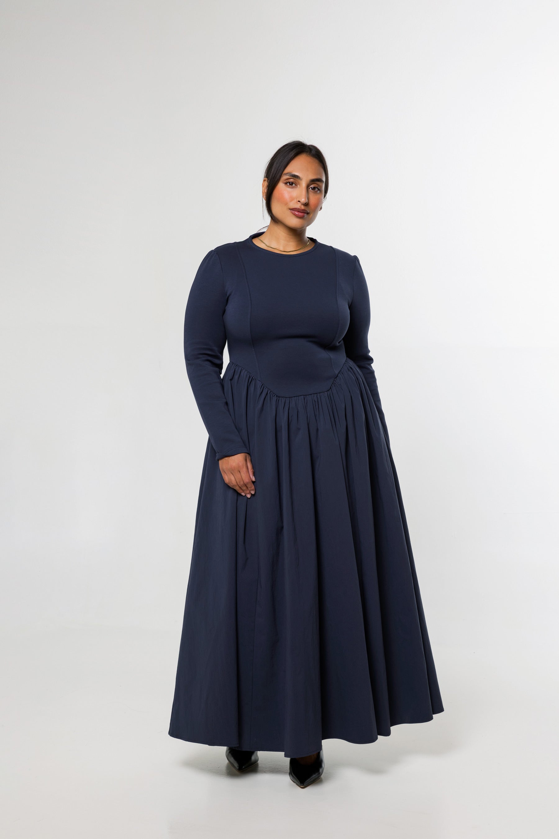 A woman modeling a long-sleeve navy blue maxi dress with a fitted bodice and a full, flowing skirt, standing against a plain light gray studio background.