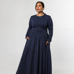 A woman modeling a long-sleeve navy blue maxi dress with a gathered waist and side pockets, standing against a light gray studio background.