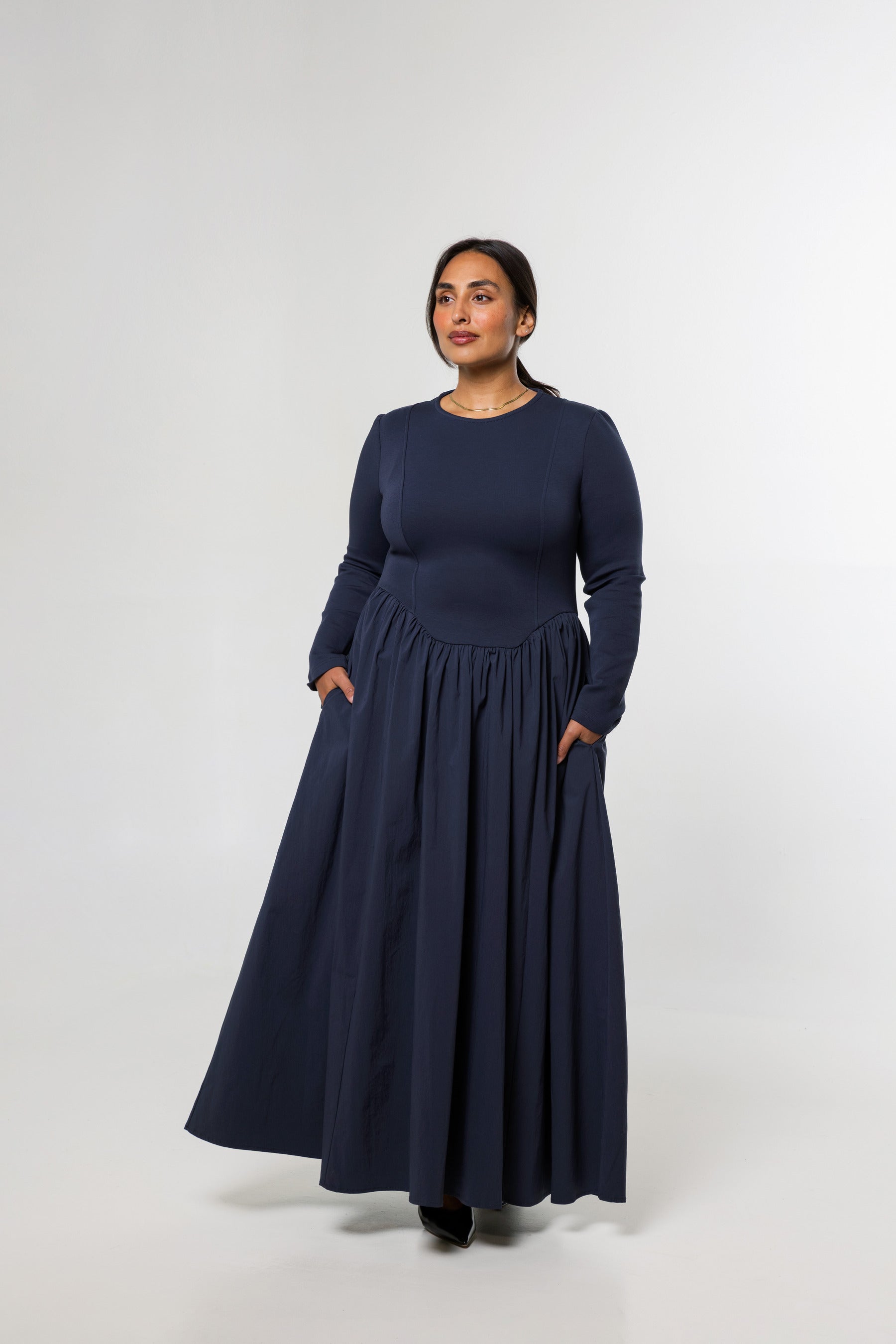 A woman modeling a long-sleeve navy blue maxi dress with a gathered waist and side pockets, standing against a light gray studio background.