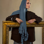 A woman wearing a slate blue ruffled hijab, a dark brown blazer, and gray trousers, posing behind a large ornate gold picture frame against a warm beige background.