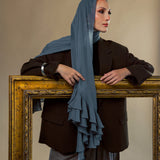 A woman wearing a slate blue ruffled hijab, a dark brown blazer, and gray trousers, posing behind a large ornate gold picture frame against a warm beige background.