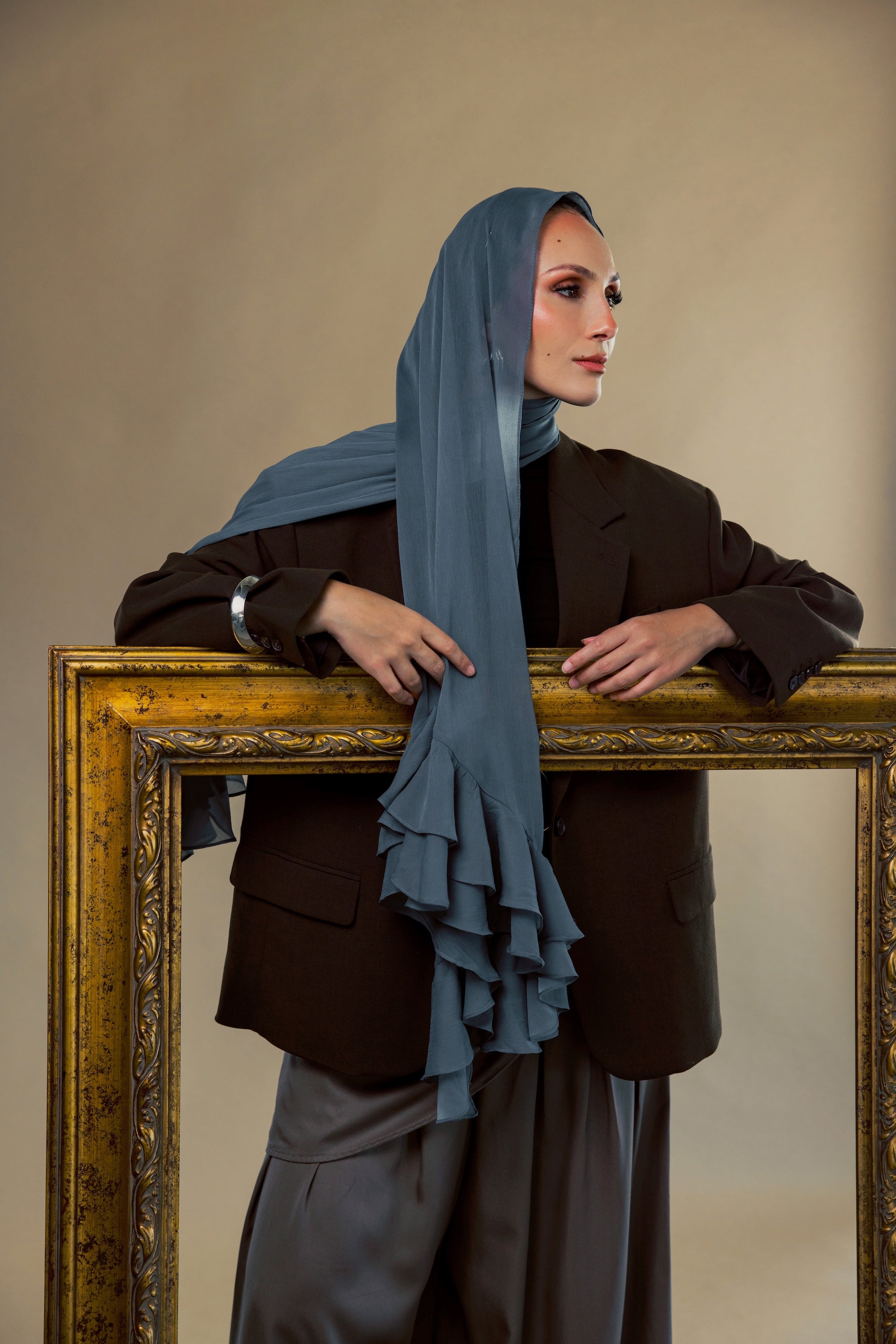A woman wearing a slate blue ruffled hijab, a dark brown blazer, and gray trousers, posing behind a large ornate gold picture frame against a warm beige background.
