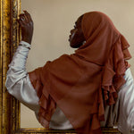 A woman in profile wearing a rust-brown ruffled hijab and a white long-sleeve top, leaning against an ornate gold-framed mirror against a beige wall.