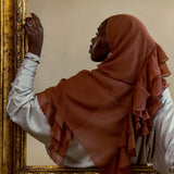 A woman in profile wearing a rust-brown ruffled hijab and a white long-sleeve top, leaning against an ornate gold-framed mirror against a beige wall.