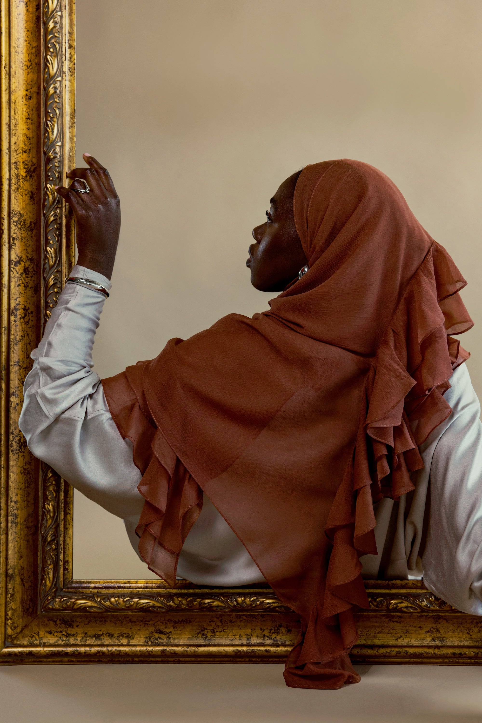 A woman in profile wearing a rust-brown ruffled hijab and a white long-sleeve top, leaning against an ornate gold-framed mirror against a beige wall.