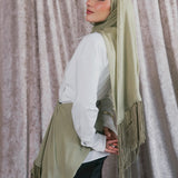 A woman wearing a sage green hijab with fringe trim, styled over a white long-sleeve blouse and black pants, standing in front of a textured gray curtain.