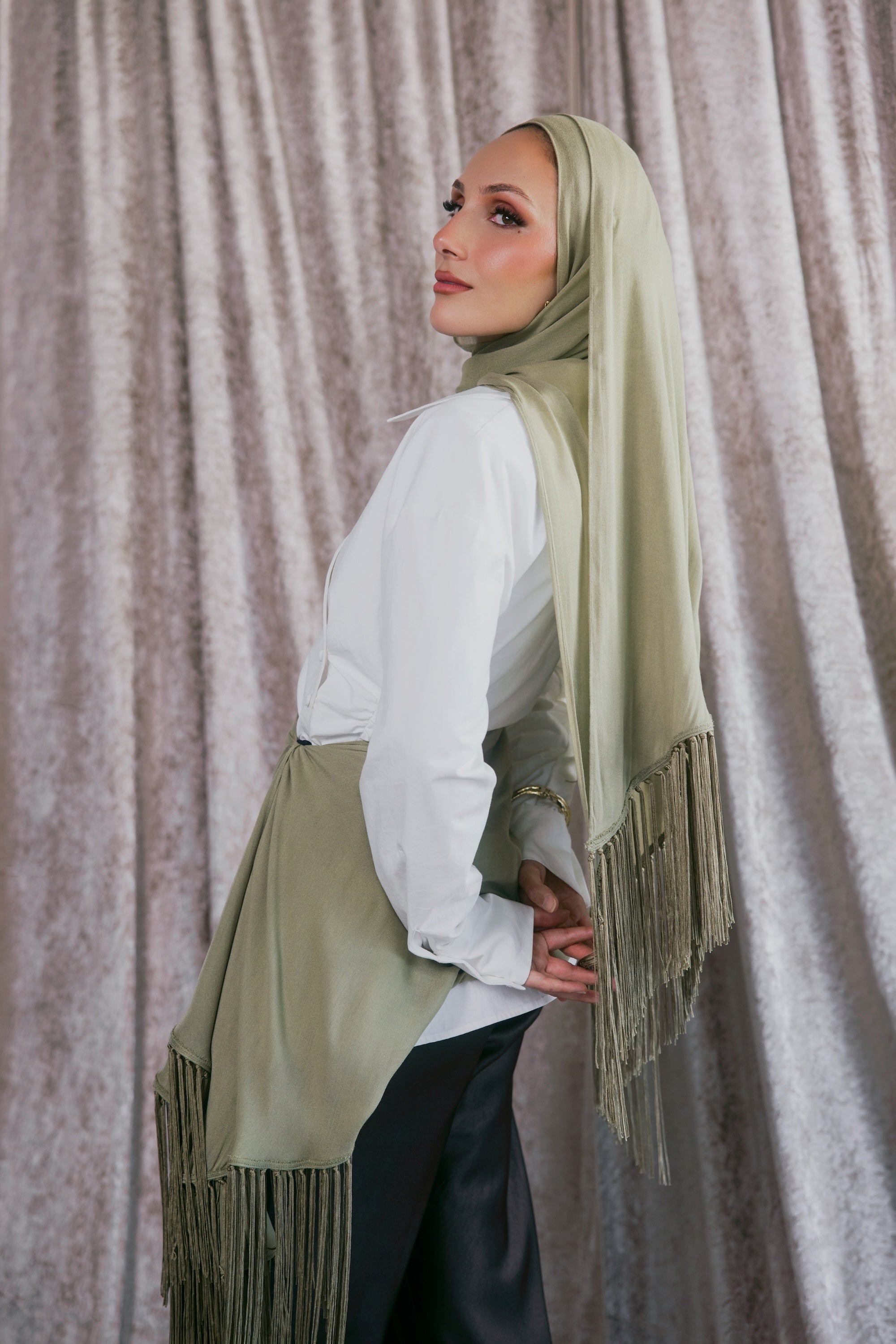 A woman wearing a sage green hijab with fringe trim, styled over a white long-sleeve blouse and black pants, standing in front of a textured gray curtain.