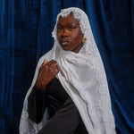 A woman wearing a white lace-trimmed shawl over a black outfit, posed against a dark blue velvet backdrop.
