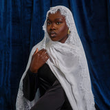 A woman wearing a white lace-trimmed shawl over a black outfit, posed against a dark blue velvet backdrop.