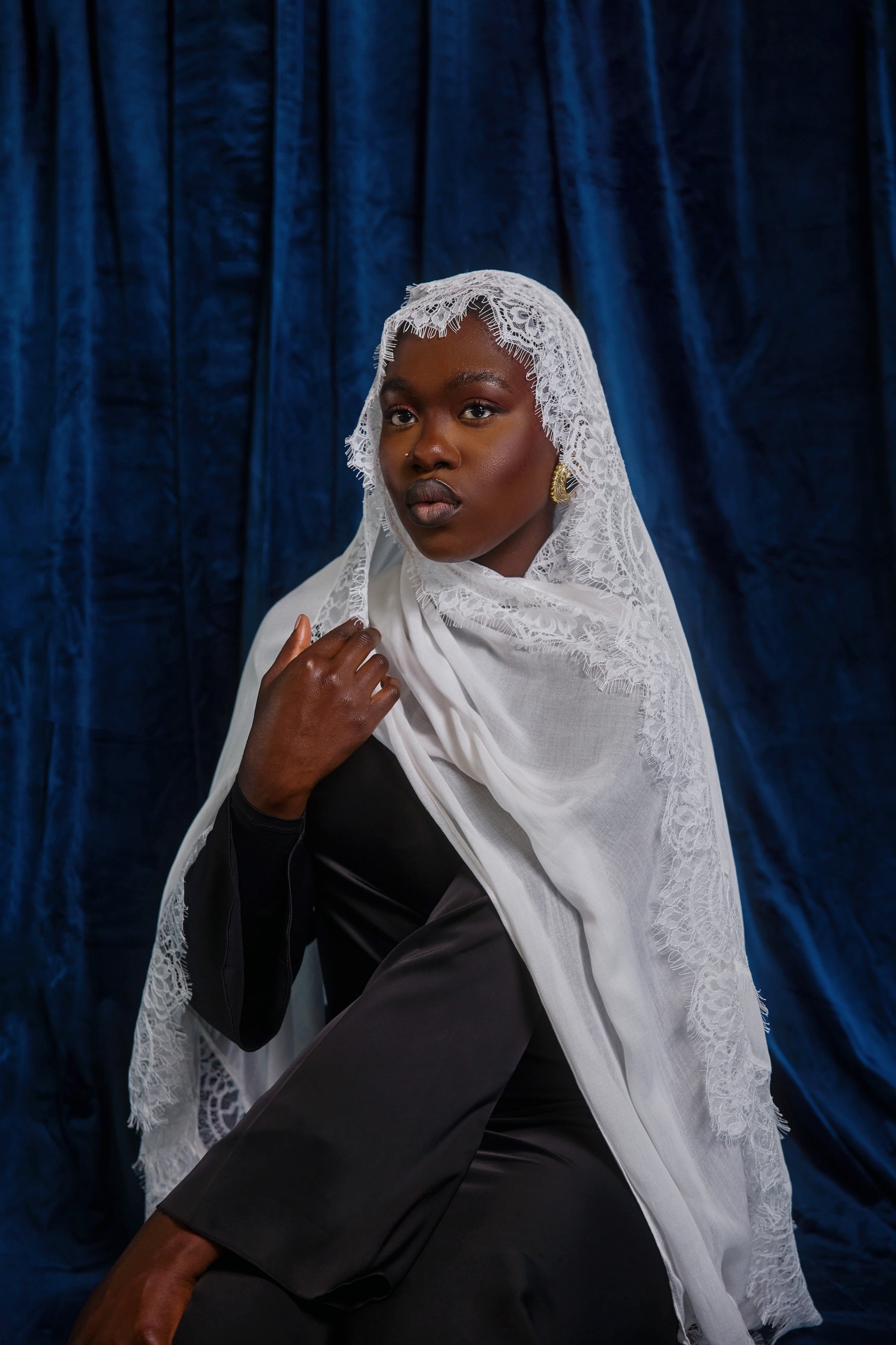 A woman wearing a white lace-trimmed shawl over a black outfit, posed against a dark blue velvet backdrop.