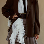 A model wearing a dark brown oversized blazer over a black top and a brown ruffled skirt, styled with a white scarf and a wide brown belt with a gold buckle, set against a solid beige background.