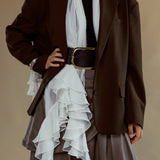A model wearing a dark brown oversized blazer over a black top and a brown ruffled skirt, styled with a white scarf and a wide brown belt with a gold buckle, set against a solid beige background.