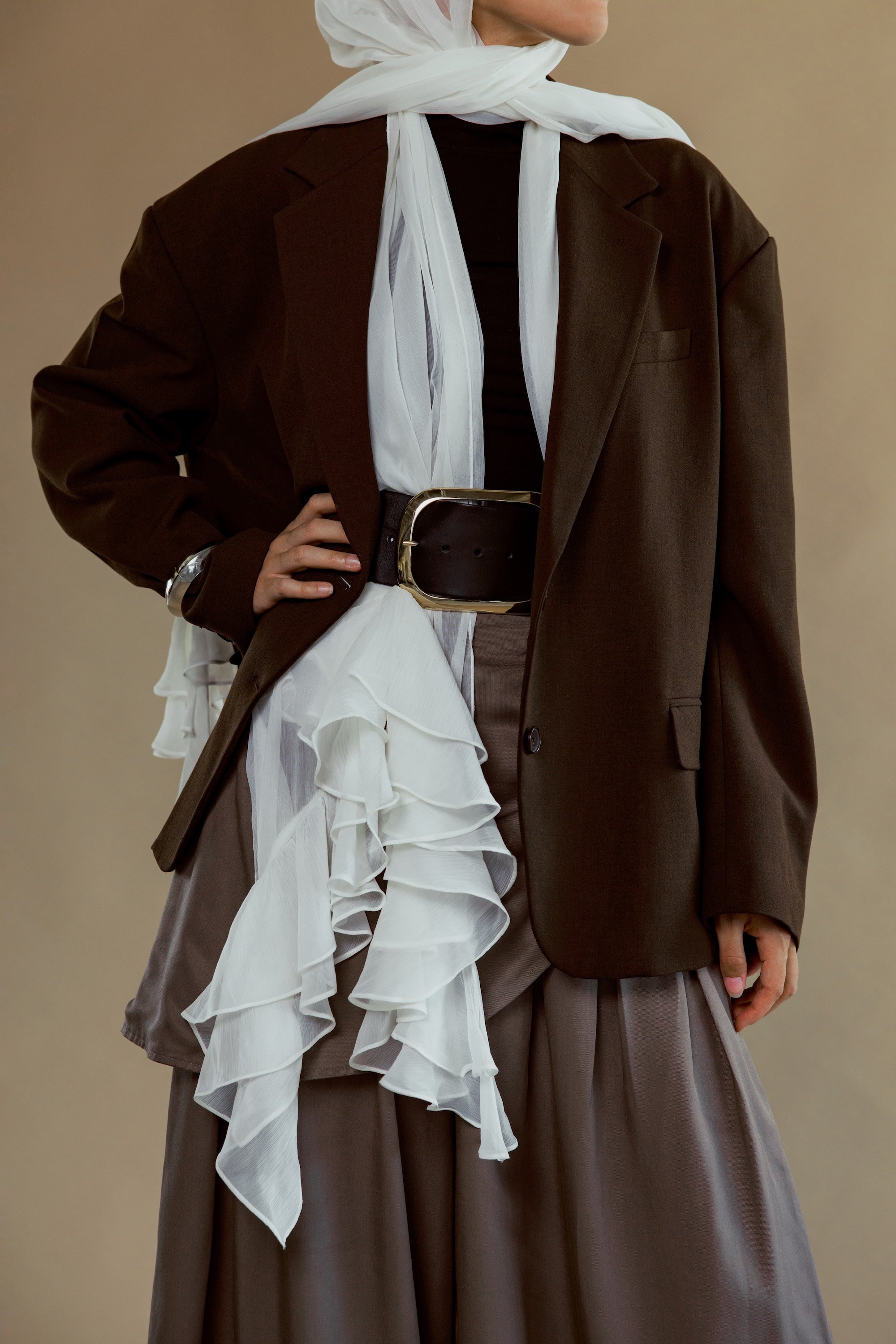 A model wearing a dark brown oversized blazer over a black top and a brown ruffled skirt, styled with a white scarf and a wide brown belt with a gold buckle, set against a solid beige background.