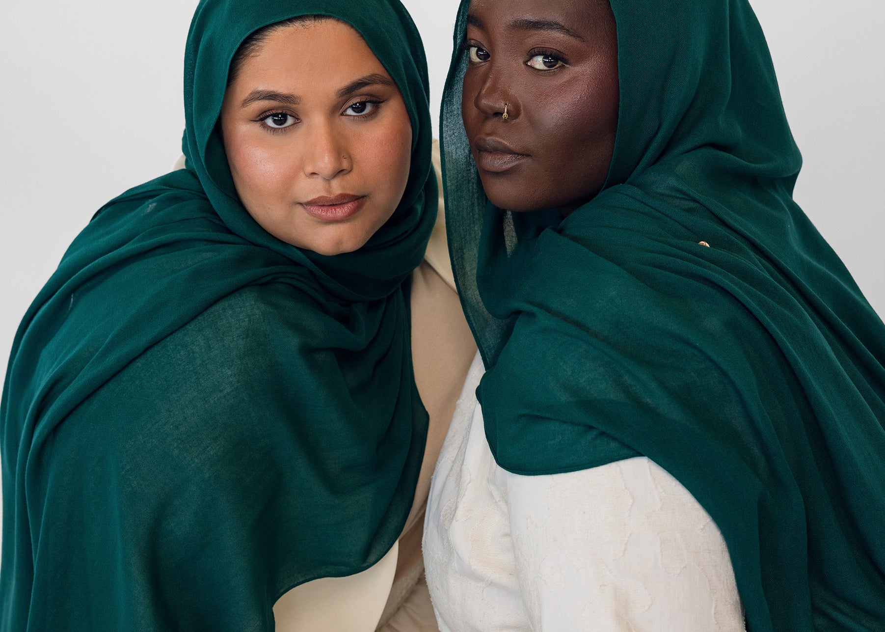 Two women wearing evergreen hijabs and white clothing against a white background