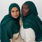 Two women wearing evergreen hijabs and white clothing against a white background